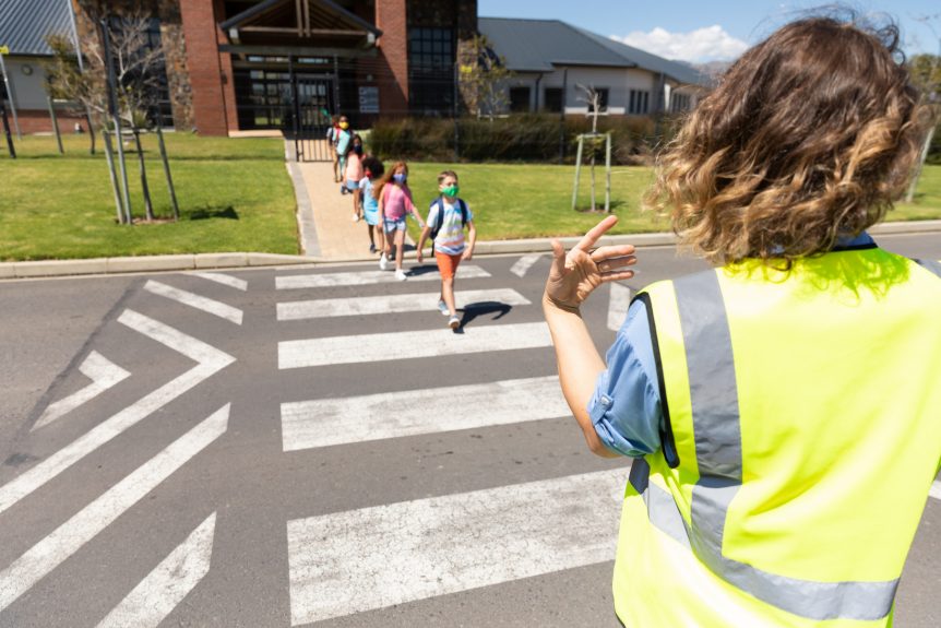 Group of children wearing face masks while they cross the road at a crossing with the help of a woman in a high vis vest