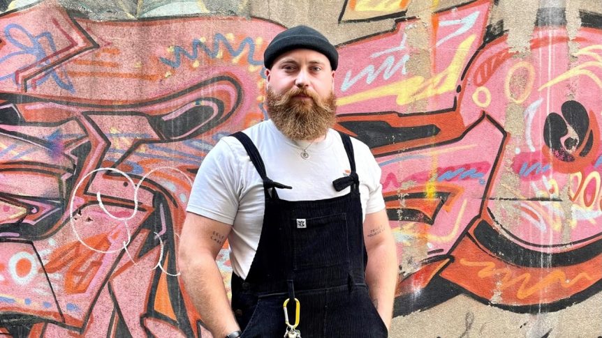 Male photography student standing in front of graffitied wall with hands in his pocket