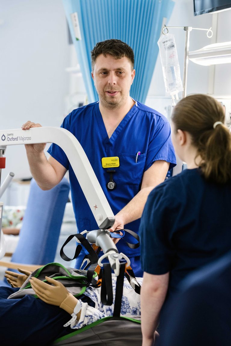 Male Nursing Associate (Higher Apprenticeship) student working in a practical setting - learning how to use a hoist with a medical dummy