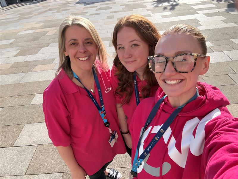 Three smiling women wearing pink tops and lanyards take a selfie outdoors on a paved walkway in bright sunlight.